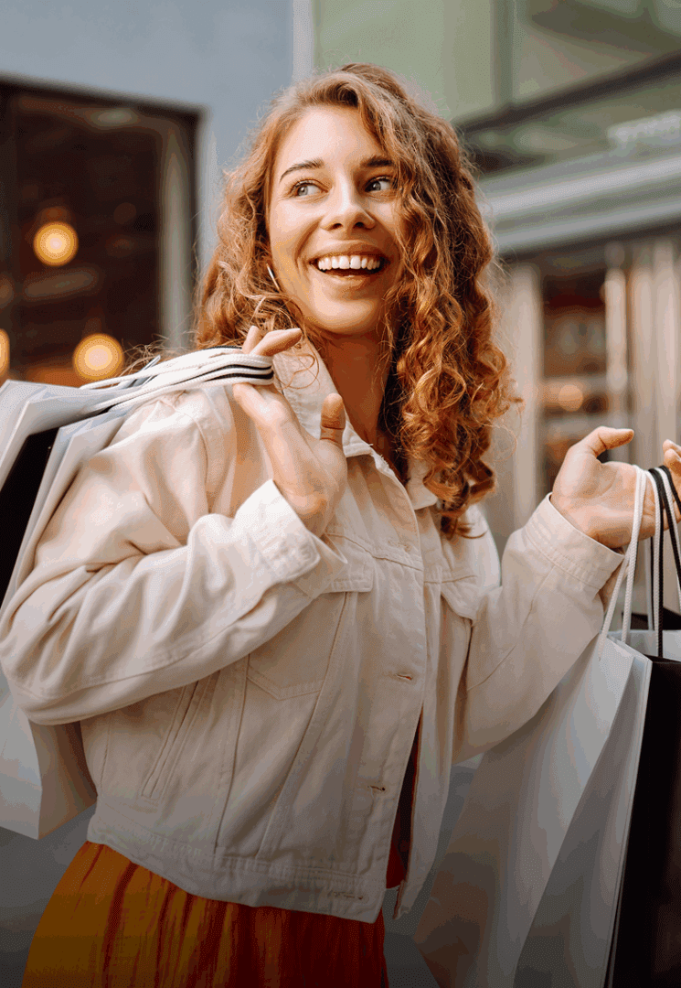 A woman with curly hair smiles while carrying multiple shopping bags over her shoulders, wearing a light jacket and an orange skirt.