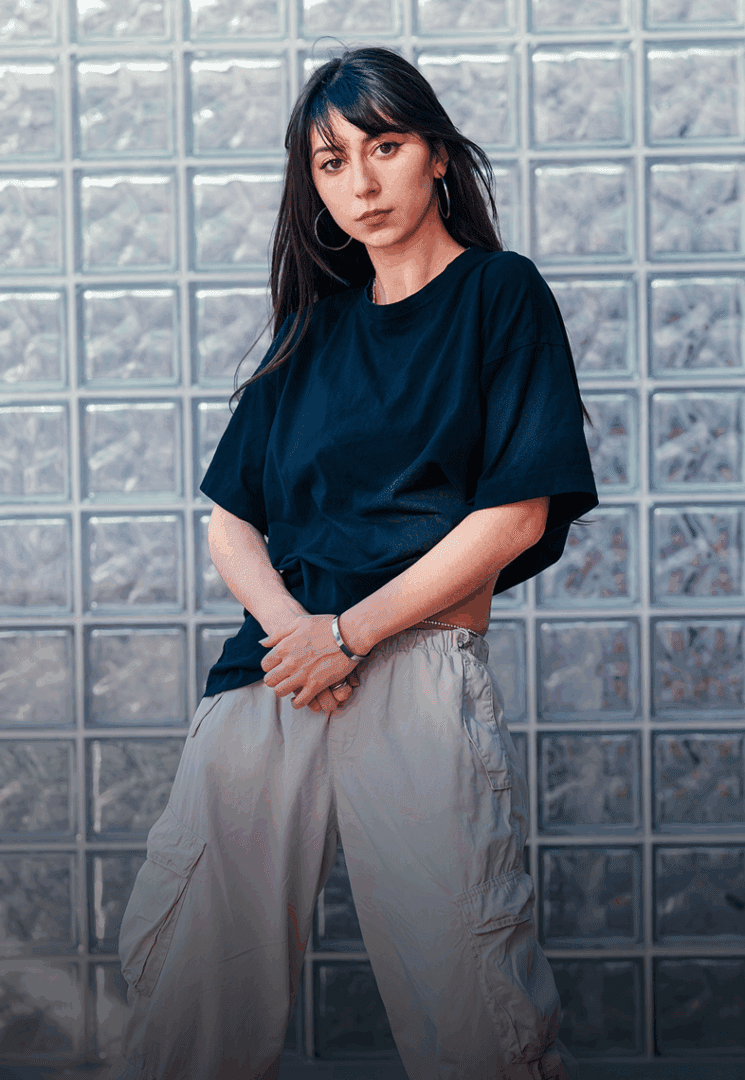 A woman with long dark hair, wearing a black T-shirt and light cargo pants, stands in front of a wall made of glass blocks.