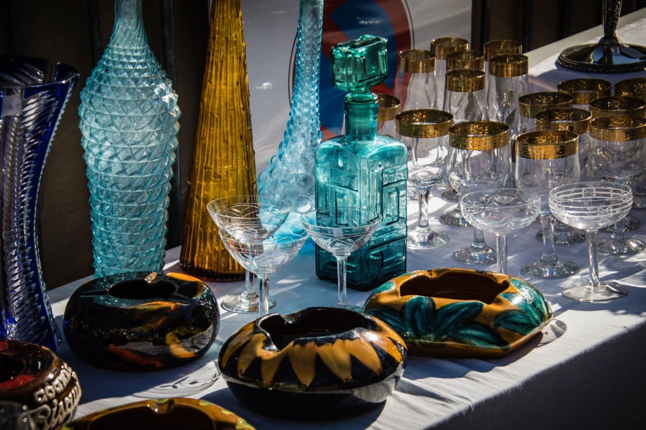 A variety of glassware and ceramic ashtrays are arranged on a white tablecloth, including colored bottles, gold-rimmed glasses, and patterned dishes.