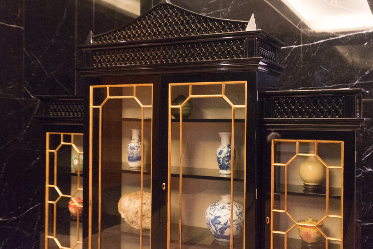A black and gold display cabinet with glass doors contains decorative vases and pottery on shelves against a marble wall background.