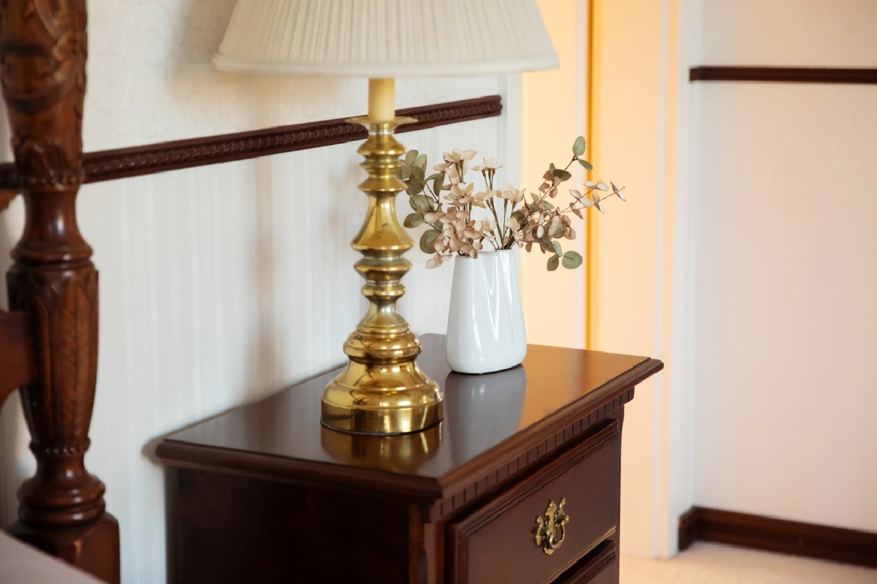 A wooden nightstand with a brass lamp and a white vase containing dried flowers, positioned against a cream-colored wall with dark trim.