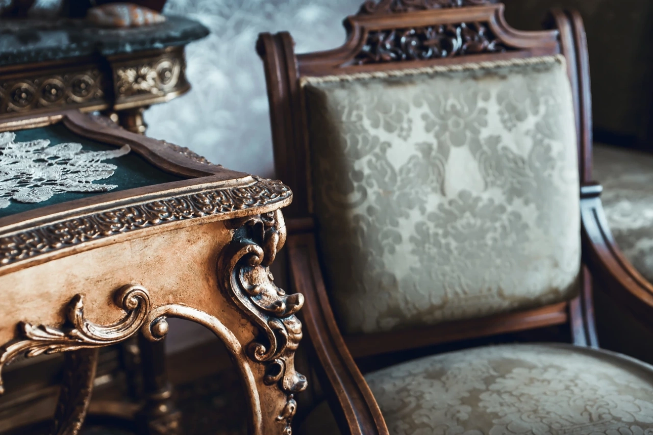 Close-up of an ornate, gold-trimmed wooden table with carved details and a lace doily beside an upholstered antique chair with patterned fabric.