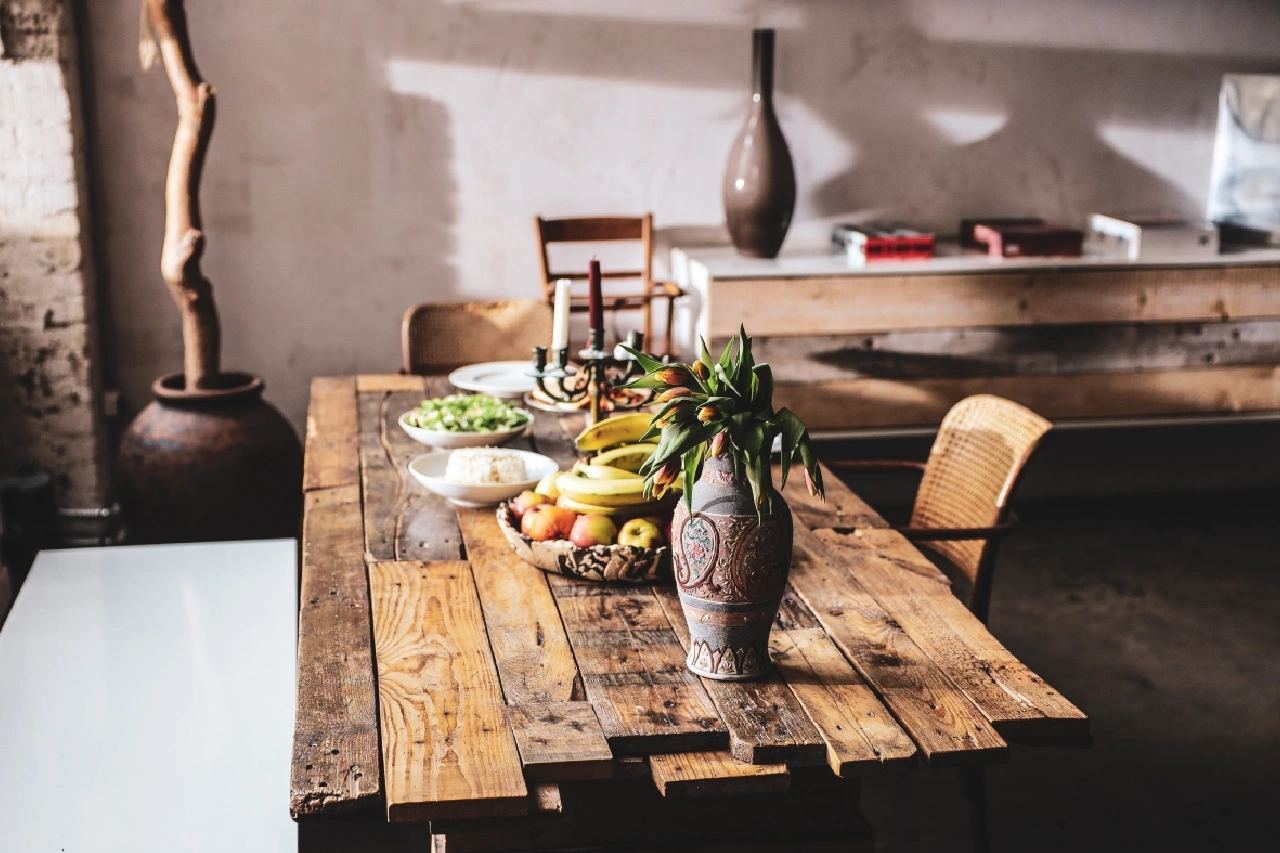 A rustic wooden dining table with a fruit bowl, candles, dishes of food, and a vase with flowers, set in a bright, minimalist room.